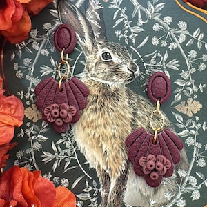 May include: A pair of dark maroon dangle earrings featuring a textured oval stud connected by a gold-toned jump ring to a fan-shaped piece with a small floral cluster at the base. The earrings are displayed against a dark green card with a rabbit illustration.