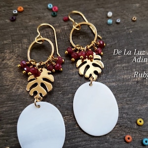 May include: Gold-tone earrings with a leaf design and white oval pendants. The earrings feature ruby-colored beads and the text "De La Luz Artisan Adina Ruby". The earrings are on a dark wood surface.