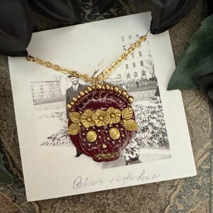 May include: A gold chain necklace with a red and gold sugar skull pendant. The pendant has gold flowers and a floral design. The necklace is displayed on a white card with the text "De La Luz Artisan" and "Primera purple Luna".