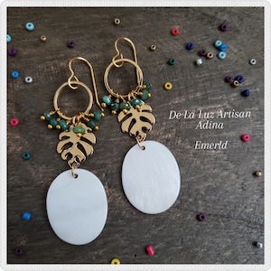 May include: Gold-tone earrings with a heart-shaped hook, a gold monstera leaf, and a white oval pendant. The earrings feature small green beads and the text "De La Luz Artisan Adina Emerld".