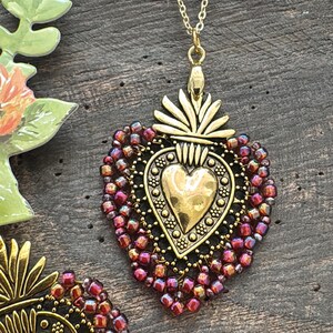 May include: A gold-toned metal heart pendant necklace featuring a central embossed heart design with a sunburst top, surrounded by a border of iridescent dark red and purple seed beads, suspended from a delicate gold-colored metal chain.
