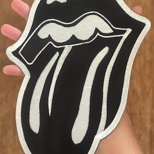 Könnte beinhalten: Schwarz-weißer, gestickter Aufnäher mit dem ikonischen Zungen- und Lippen-Logo der Rolling Stones. Der Aufnäher hat einen schwarzen Hintergrund mit weißen Stickdetails und einem weißen Rand. Das Design ist eine stilisierte Darstellung eines Mundes mit herausgestreckter Zunge.