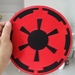 Può includere: Una toppa ricamata circolare con un cerchio centrale nero e sei raggi, circondata da un campo rosso e un anello esterno nero con un bordo geometrico dentellato. Il disegno è realizzato con filo di colore rosso e nero brillante.