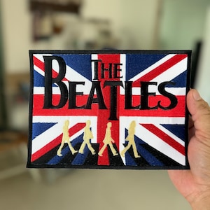 Könnte beinhalten: Ein gestickter Aufnäher mit der Union Jack Flagge mit den Worten "The Beatles" in schwarzer Schrift und vier Figuren, die in Gold über die Flagge laufen.