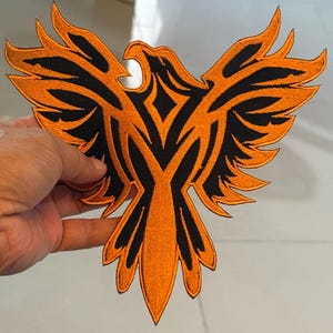 May include: Embroidered patch depicting a phoenix with spread wings. The patch has an orange background with black detailing, creating a striking contrast. The patch is likely for clothing or accessories.