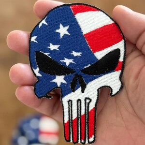 May include: A white skull with a red, white, and blue American flag design. The skull is outlined in black and has a black eye socket.