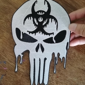 May include: A white and black embroidered patch featuring a stylized skull design. The forehead area displays a black biohazard symbol, while the eye sockets and nasal cavity are black. The lower portion of the skull appears to be melting.