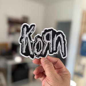 以下が含まれることがあります： 「Korn」という単語がスタイリッシュなフォントで刺繍された黒と白の刺繍パッチ