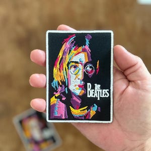 Könnte beinhalten: Ein farbenfroher gestickter Patch mit einem Porträt von John Lennon und dem Text "The Beatles" in weißen Buchstaben. Der Patch ist auf schwarzem Hintergrund mit weißem Rand.