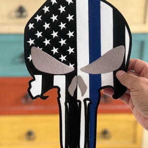 May include: A black and white skull with an American flag and a blue line on the right side. The skull is outlined in black and has a white background.