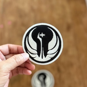 May include: A white and black embroidered patch featuring the symbol of the Mandalorian faction from the Star Wars universe. The symbol is a stylised image of a creature with wings and a central crest.