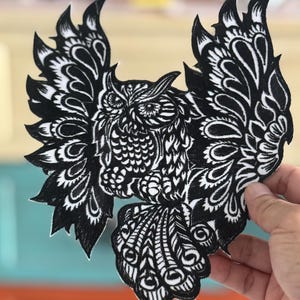 May include: Black and white embroidered patch of an owl with its wings spread out. The owl has intricate details and a detailed design.