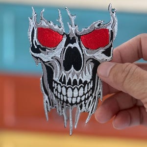 Peut inclure: Un patch brodé de crâne blanc et gris avec des yeux rouges. Le crâne a un effet fondu qui coule.