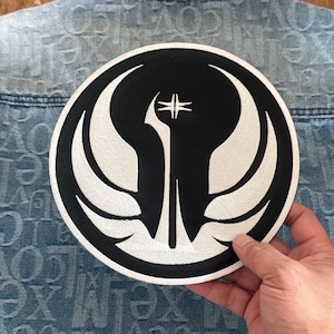 May include: A white and black embroidered patch with a stylized symbol of a winged creature. The patch is circular and has a white outline.