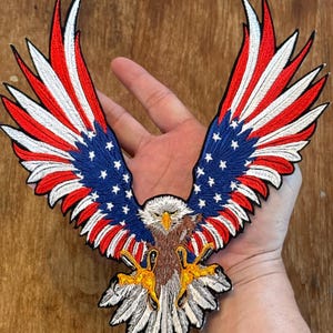 May include: An embroidered patch of a bald eagle with its wings spread wide. The eagle's wings are patterned with the American flag, with red and white stripes and blue stars. The eagle is brown and white with yellow talons.