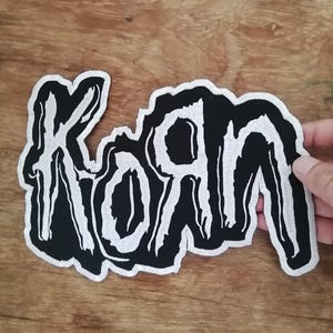 May include: A black and white embroidered patch featuring the band logo Korn, with the letters stylized in a jagged, distressed font. The design consists of white lettering outlined in black, creating a high-contrast graphic suitable for application.