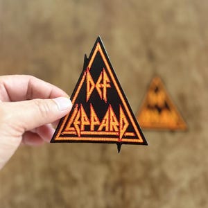以下が含まれることがあります： オレンジ色の刺繍で「Def Leppard」ロゴが施された黒い三角形のワッペンです。