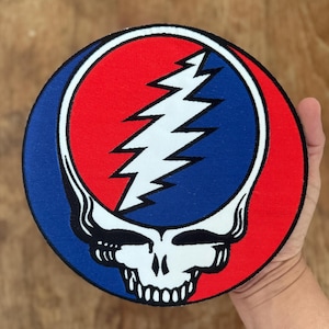 Puede incluir: Un parche redondo rojo, blanco y azul con un diseño de calavera y rayo. La calavera es blanca con ojos negros y una boca negra. El rayo es blanco y está dentro de un círculo azul. El círculo está rodeado por un anillo rojo.