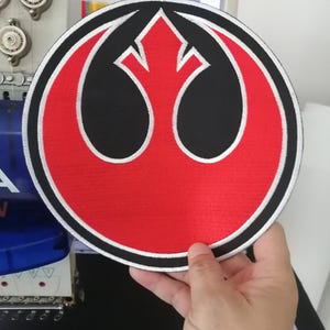 May include: A circular embroidered patch featuring a red and black Rebel Alliance symbol from Star Wars. The design consists of a central red stylised phoenix emblem set against a black background, enclosed by a thick white and black border.