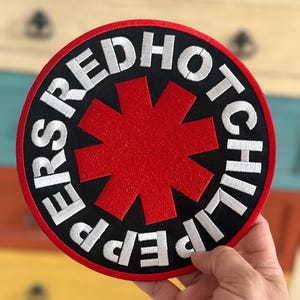 Op de afbeelding: Red Hot Chili Peppers bandlogo patch met een rode sterburst op een zwarte achtergrond en witte letters rond de rand.