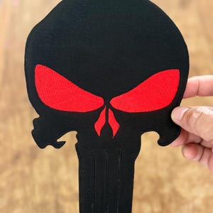 Puede incluir: Parche de calavera bordado en negro y rojo con contorno blanco. La calavera tiene una forma de calavera clásica con una mandíbula prominente y una nariz grande y prominente. Los ojos son rojos y los dientes son blancos.