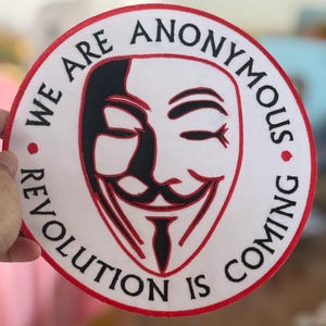 May include: A white round patch with a red border. The patch features a black and red stylized image of the Guy Fawkes mask with the text "We are Anonymous. Revolution is coming." in black.