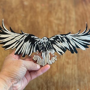May include: Black and white embroidered iron-on patch of an eagle with its wings spread wide. The eagle has a brown beak and talons.