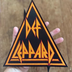 May include: A black triangular patch with an orange embroidered outline. The patch features the text "Def Leppard" in a stylized font.