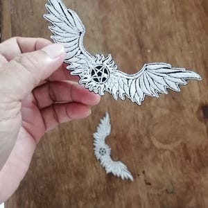 May include: An embroidered patch featuring white wings extending from a central circular emblem containing a black pentagram. The design is outlined in black thread, creating a high-contrast graphic element suitable for application onto fabric surfaces.