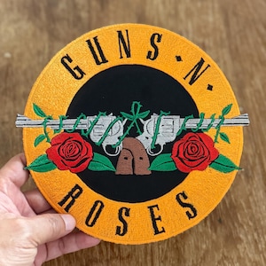 Puede incluir: Un parche bordado amarillo y negro con el logotipo de Guns N' Roses. El logotipo presenta dos pistolas cruzadas con rosas y espinas, rodeado de un círculo amarillo con el nombre de la banda.