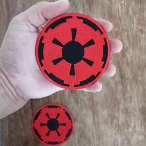 Può includere: Due toppe ricamate circolari con sfondo rosso, un emblema centrale nero simile a un sole e un design a cerchio esterno nero. La toppa più grande è tenuta nel palmo della mano, mentre la più piccola è posizionata al di sotto.