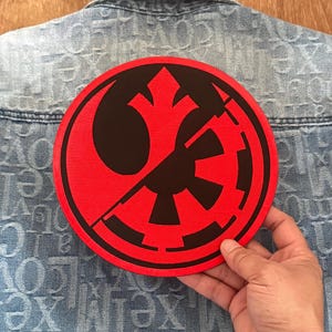 Könnte beinhalten: Ein runder roter und schwarzer Patch mit dem Symbol der Rebellenallianz auf der linken Seite und dem imperialen Symbol auf der rechten Seite. Die Symbole sind in der Mitte geteilt, wodurch ein kombiniertes Design entsteht.