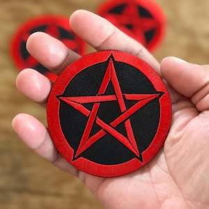 Può includere: Una toppa ricamata con un pentagramma rosso su sfondo nero e un cerchio esterno rosso. La toppa è tenuta in una mano.