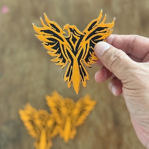 May include: Embroidered patch featuring a stylized phoenix design. The patch has a vibrant orange and black color scheme, with the phoenix depicted in flight. The patch is held up by a hand, with two additional patches visible in the background.