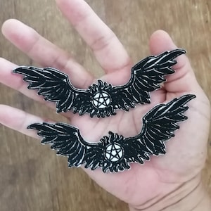 Puede incluir: Dos parches bordados negros con alas estilizadas que se extienden desde un emblema circular central. Cada emblema contiene un pentagrama blanco rodeado por un borde negro en forma de llama. Los parches tienen costuras detalladas.