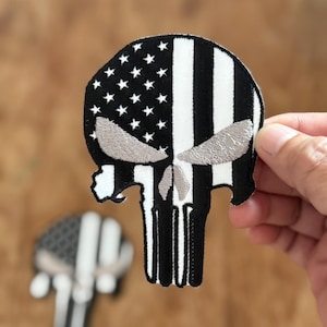 May include: Black and white embroidered patch of a skull with an American flag design. The skull has a white outline and the flag is split down the middle with the stars on the left and the stripes on the right.