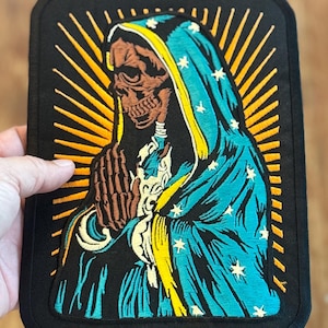 May include: Embroidered patch featuring a skeletal figure in a blue and yellow robe with a halo of yellow rays. The figure has a skull face and is holding its hands in a prayer position.