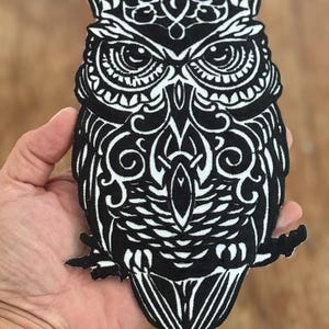 May include: Black and white embroidered iron-on patch of an owl with intricate details. The owl is perched on a branch with its wings spread out.