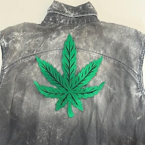 May include: A sleeveless denim jacket in a faded grey wash. A large, embroidered green cannabis leaf is centered on the back. The jacket has a classic collar and seams.