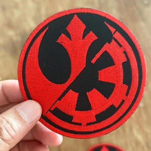Op de afbeelding: Een rood en zwart geborduurd patch met het Rebel Alliance-symbool aan de linkerkant en het Imperiale symbool aan de rechterkant, beide symbolen overlappen elkaar gedeeltelijk.