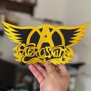 Peut inclure: Un patch brodé or avec le logo d'Aerosmith. Le logo représente un 'A' stylisé avec des ailes et le nom du groupe écrit en écriture cursive.
