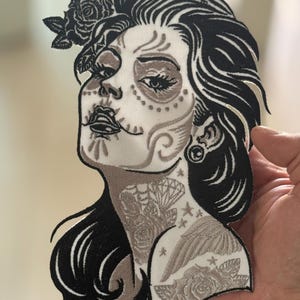 Puede incluir: Parche bordado para planchar con una ilustración detallada de una mujer con un diseño de maquillaje de calavera de azúcar. La mujer tiene el pelo largo y negro, una corona de flores y tatuajes intrincados en el cuello y el hombro. El parche está en tonos negros y grises.