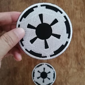 Op de afbeelding: Twee ronde geborduurde patches met het zwart-witte symbool van het Galactische Keizerrijk. Het ontwerp bestaat uit een centrale zwarte cirkel met zes stralen, omringd door een zwart-witte geometrische rand met een gestikte afwerking.