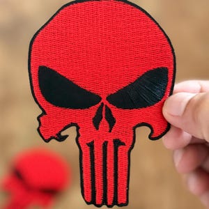 Könnte beinhalten: Roter gestickter Schädel-Patch mit schwarzen Augen und einem weißen Schädel-Umriss. Der Schädel ist ein beliebtes Symbol des Punishers.