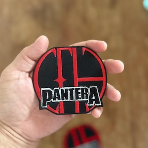 Puede incluir: Parche bordado en negro y rojo con el nombre de la banda "PANTERA" en letras blancas. El logotipo de la banda es un diseño estilizado con líneas rojas y un borde circular. El parche se sostiene en una mano.
