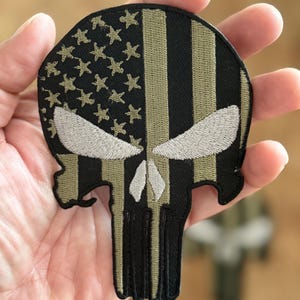 Puede incluir: Un parche bordado negro y verde del cráneo del Punisher con un diseño de bandera estadounidense. El cráneo está contorneado en blanco y la bandera es de color verde oliva y negro.