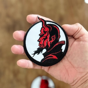 Può includere: Toppa ricamata in rosso e nero di una testa di diavolo di profilo, con corna e barba, su uno sfondo circolare bianco.
