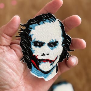 Pode incluir: Etiqueta bordada do Joker do filme O Cavaleiro das Trevas. A etiqueta apresenta um close-up do rosto do Joker com um fundo branco, cabelo preto e um sorriso vermelho e azul.