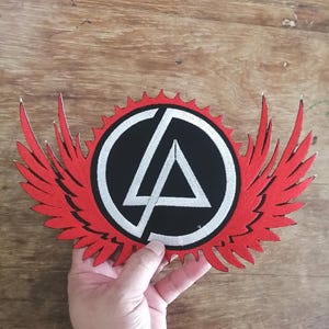May include: Embroidered patch featuring a black circle with a white triangle and lines, surrounded by a red sunburst design and red wings. The patch is held up against a wooden surface.
