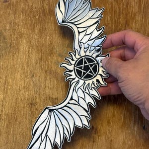 Peut inclure: Patch brodé blanc avec un pentagramme noir à l'intérieur d'un cercle entouré de flammes. Le patch est en forme d'ailes d'ange.